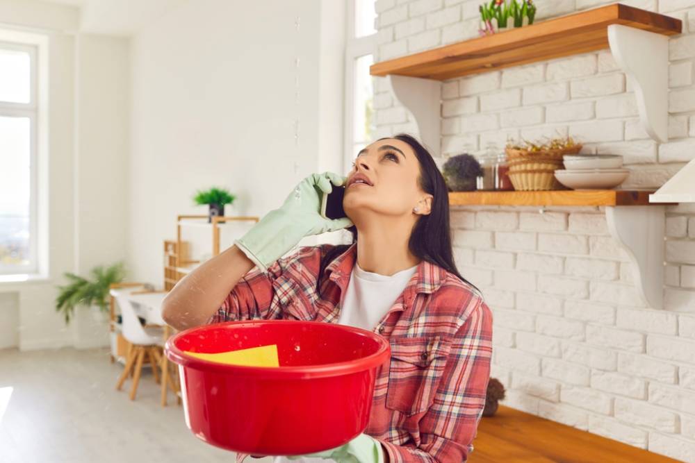 Having a roof leak at home. Worried young woman in rubber gloves holding red plastic basin
